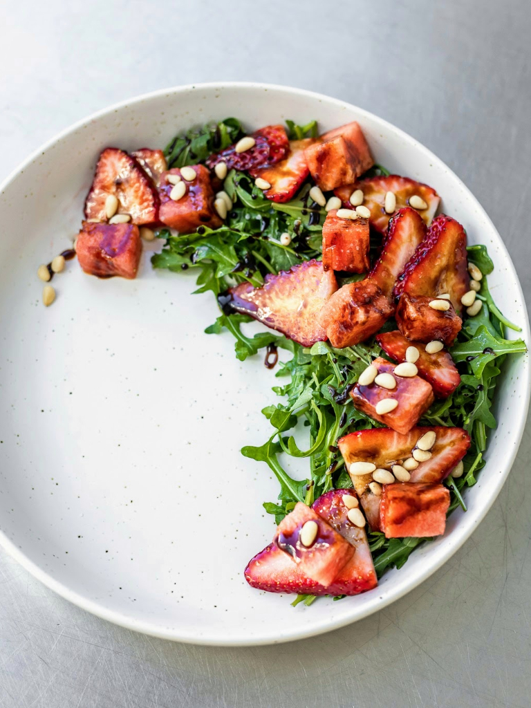 A plate with strawberries, arugula and pine nuts.